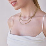 Close-up of a woman's décolletage wearing a Bold Link Chain and a Chic Cuban Curb Chain, both in yellow gold, with a white strap top against a light grey background.
