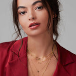 A brunette woman wearing several yellow gold necklaces - a Straight as an Arrow Necklace, a Pavé Horizon Necklace, a Custom Gold Necklace - 5 Letters with birthstones, a LOVE Necklace, and a Gold Lariat Necklace, along with Tres Pearl Earrings, with a silk red top against a light grey background.