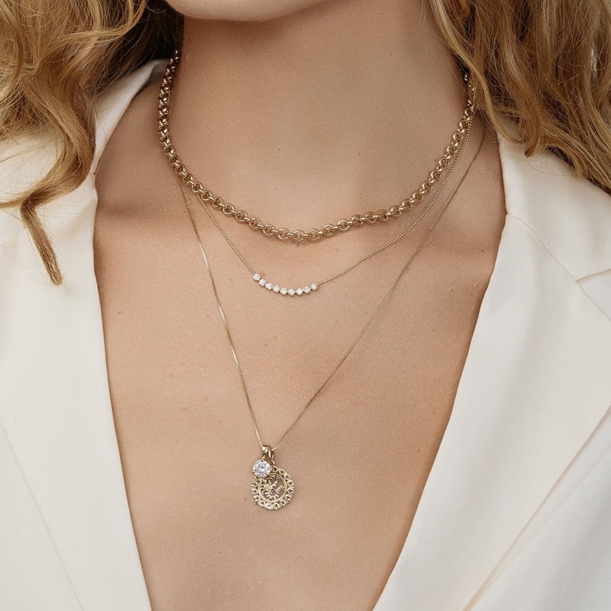 Close-up of a blonde woman's décolletage wearing a Rolo Necklace, a Diamond Arc Layering Necklace and a Zodiac Necklace - Scorpio with a Princess Diamond Pendant, yellow gold, with a white top. 