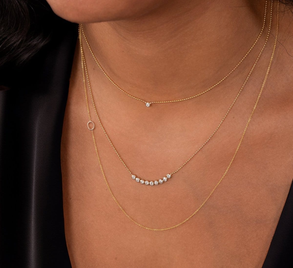 Close-up of a neck wearing a Single Diamond Layering Necklace, a Diamond Arc Layering Necklace and a 14k Gold Asymmetrical Letter Necklace - O all in yellow gold, with a black silk top. 