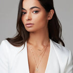 A brunette woman wearing layered necklaces - a Pavé Horizon Necklace, a Gold Lariat Necklace, and a Zodiac Necklace - Leo, along with a 14K Gold Letter Stud - M, all in yellow gold, with a white blazer against a light grey background.