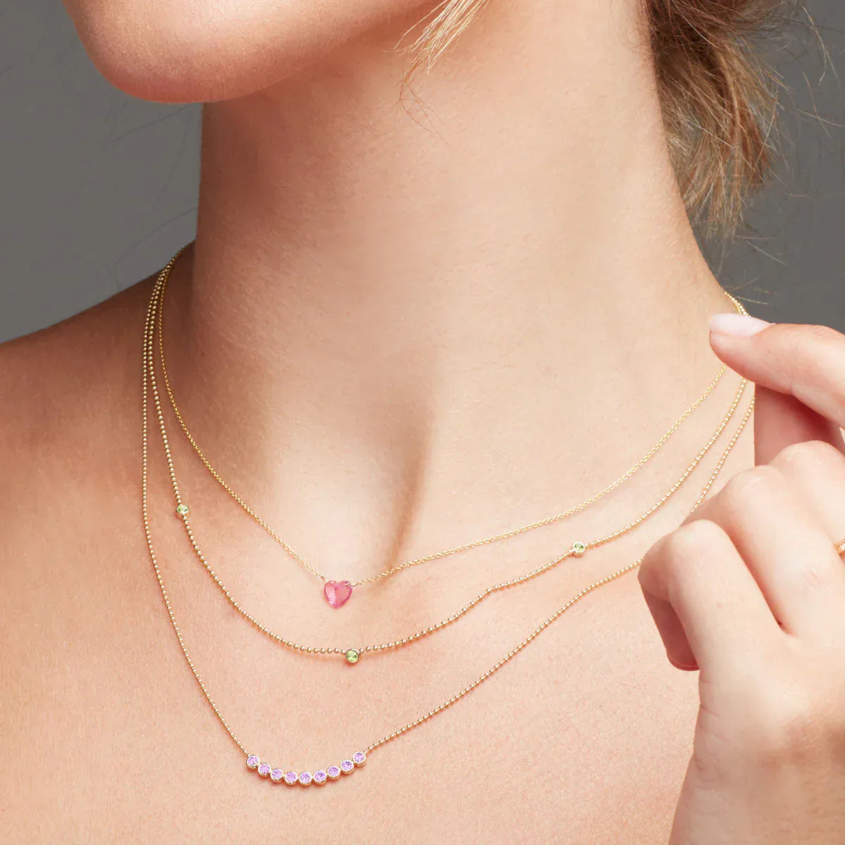 Close-up of a neck wearing The Sweetheart Necklace - Pink Sapphire, a Triple Birthstone Layering Necklace and a Birthstone Arc Layering Necklace, all in yellow gold, against a grey background. 