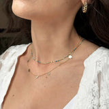 A close-up of a woman’s neck area wearing a Custom Gold Necklace in yellow gold with six charms (a star, heart, moon, birthstone, paw and letter O), layered with a Confetti Necklace in blue and Link Hoops.