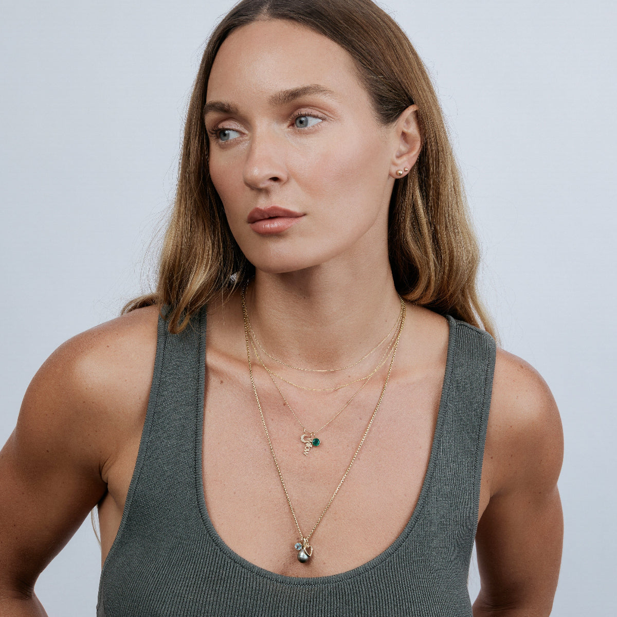 A woman wearing two Signature Cable Chains, a Classic Bead Chain with a Color Pop Pendant in Green (Emerald), a Petite Nameplate Pendant and Pavé Moon Charm, a Box Chain featuring a Color Pop Pendant in Light Blue (Aquamarine), Cut-Out Heart Charm and Baroque Pearl Pendant, and Preppy Post and Mini Preppy Post earrings, all in yellow gold, wearing a grey top with blonde hair against a light grey background.