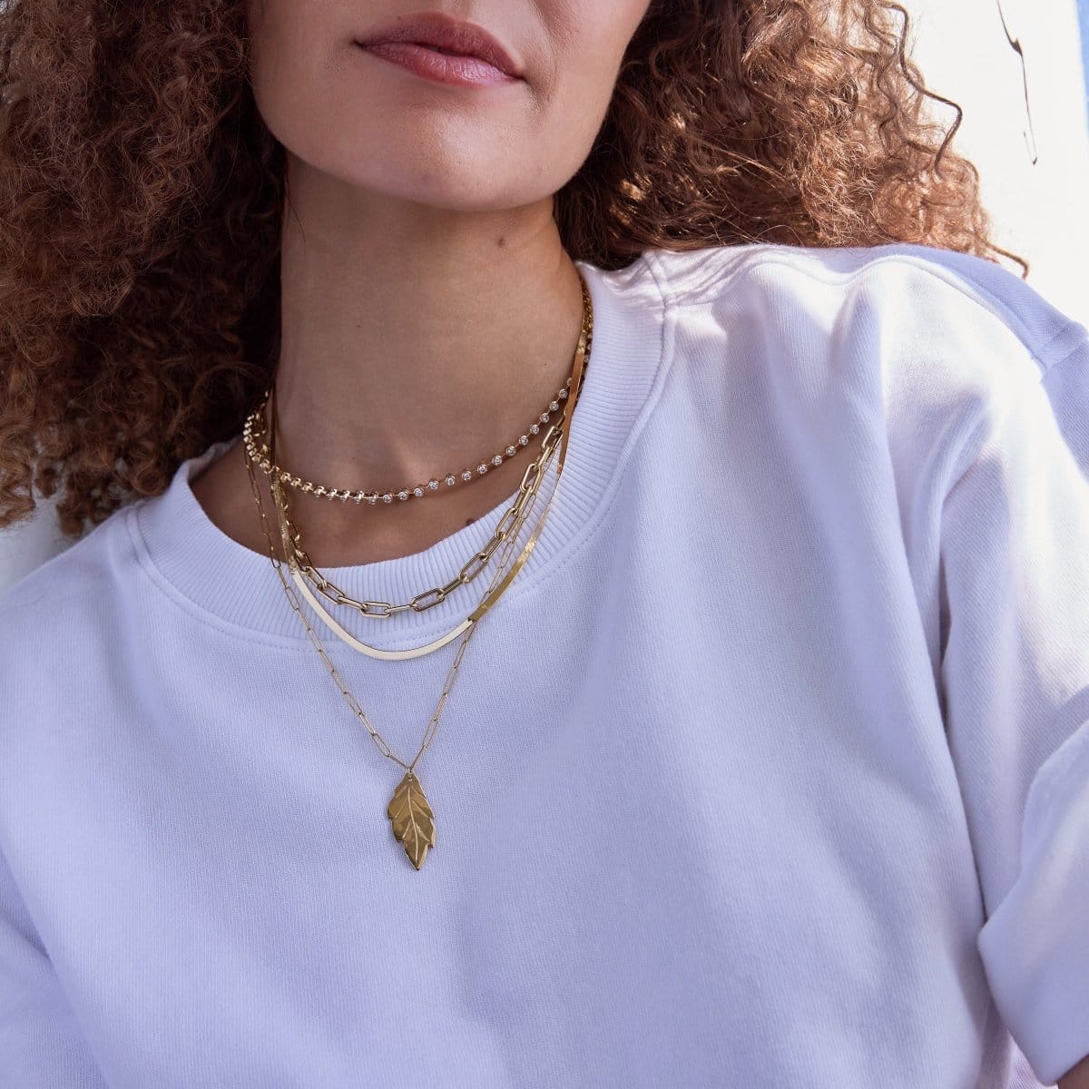 Close-up of a woman's neck wearing The Ultimate Diamond Necklace, a Bold Link Chain, Sequence Chain and Element Long Link Chain with a 20th Anniversary Leaf Pendant, styled with a white T-shirt and her brown curly hair visible.