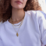 Close-up of a woman's neck wearing The Ultimate Diamond Necklace, a Bold Link Chain, Sequence Chain and Element Long Link Chain with a 20th Anniversary Leaf Pendant, styled with a white T-shirt and her brown curly hair visible.