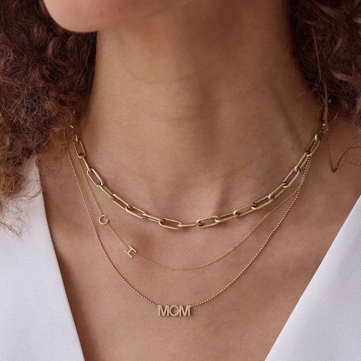 Close-up of a woman's neck wearing a Bold Link Chain, a Custom Gold Necklace — 2 Letters (C and H) and a Pavé MOM Necklace, all in yellow gold, with a white top and brown curly hair visible.