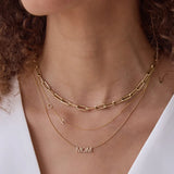 Close-up of a woman's neck wearing a Bold Link Chain, a Custom Gold Necklace — 2 Letters (C and H) and a Pavé MOM Necklace, all in yellow gold, with a white top and brown curly hair visible.