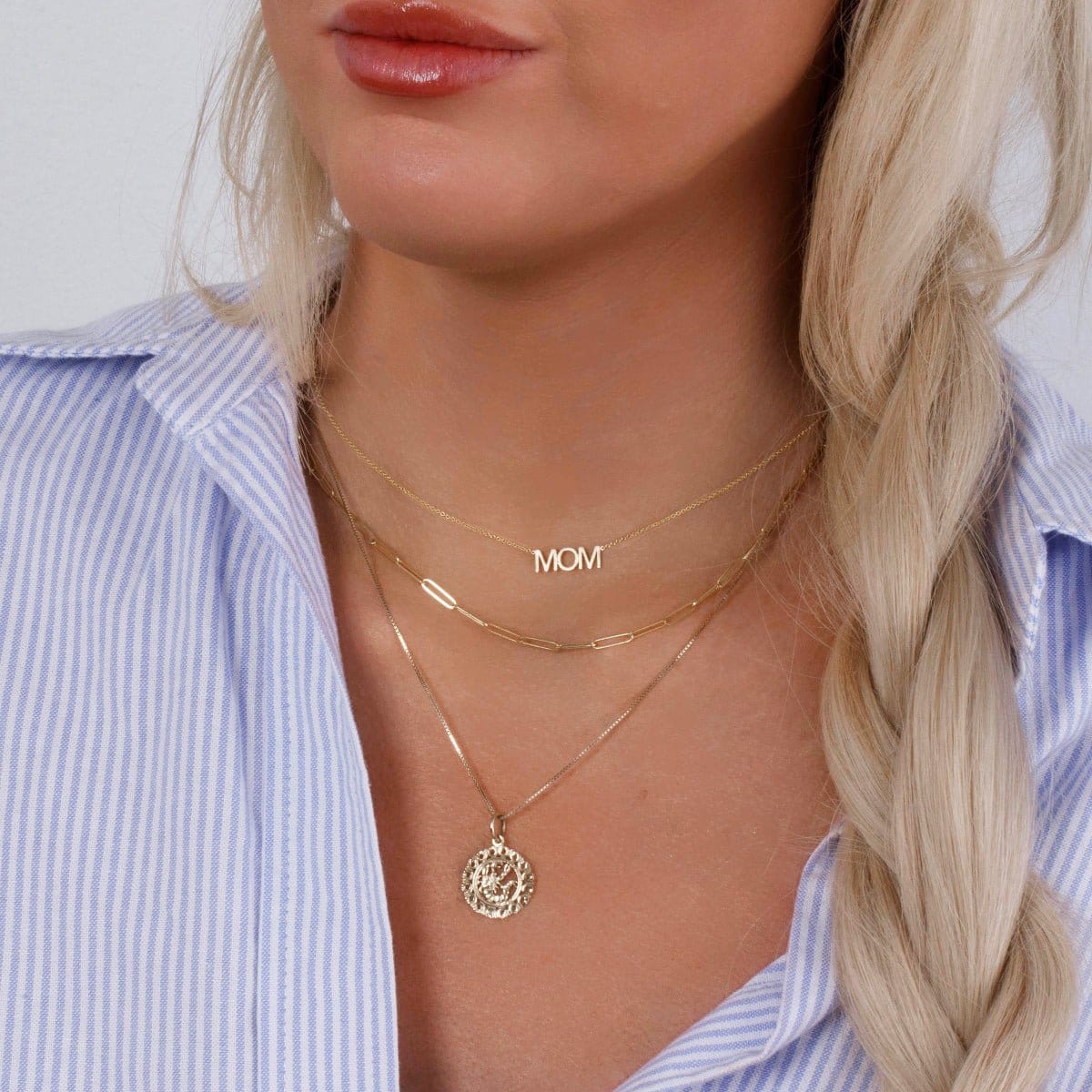 Close-up of a blonde woman's décolletage wearing a MOM Necklace, an Element Long Link Chain and a Zodiac Necklace - Scorpio, all in yellow gold, with a blue and white striped shirt against a light grey background.