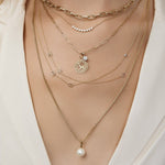 A close-up of a neck wearing a Bold Link Chain, a Diamond Arc Layering Necklace, an Element Long Link Chain with a Diamond Pendant and Zodiac Pendant (Aries) and two Custom Gold Necklaces - 3 Letters, and a Box Chain with a Perla Pendant, all in yellow gold, styled with a cream top. 