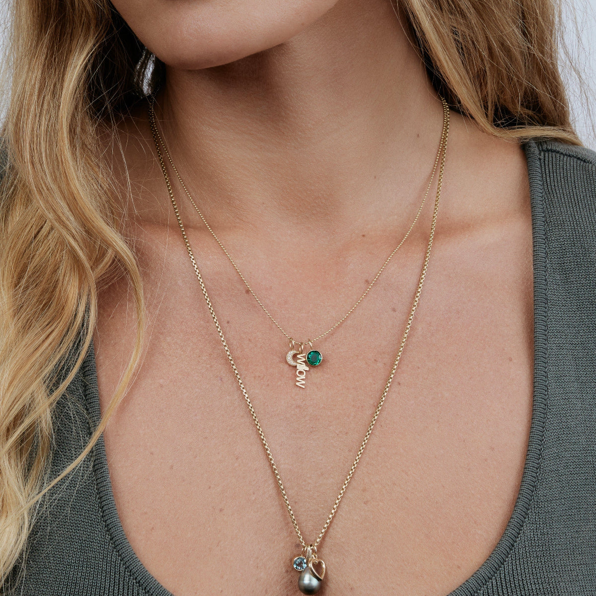 Close-up of a woman wearing a  a Classic Bead Chain with a Color Pop Pendant in Green (Emerald), a Petite Nameplate Pendant and Pavé Moon Charm, and a Box Chain with a Color Pop Pendant in Light Blue (Aquamarine), Cut-Out Heart Charm and Baroque Pearl Pendant, wearing a grey top with her blonde hair visible. 
