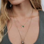 Close-up of a woman wearing a  a Classic Bead Chain with a Color Pop Pendant in Green (Emerald), a Petite Nameplate Pendant and Pavé Moon Charm, and a Box Chain with a Color Pop Pendant in Light Blue (Aquamarine), Cut-Out Heart Charm and Baroque Pearl Pendant, wearing a grey top with her blonde hair visible. 