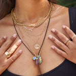 Close-up of a woman's décolletage wearing an Element Long Link Chain, a Rolo Necklace, an Overboard Layering Necklace, a Custom Gold Necklace - 4 Letters, a Single Diamond Layering Necklace, a Zodiac Necklace - Gemini, a Black Rhodium Antique Bead Chain and a Box Chain with a Balance Crystal Charm, a Small Abalone Cabochon Charm, a Diamond Heart Locket Charm, an Opal Dangle Charm, an Etched Turquoise Heart charm, a Thick Band, Delicate Stacking Trio Rings, Pave Diamond Rings and a Pave Geo Ring.
