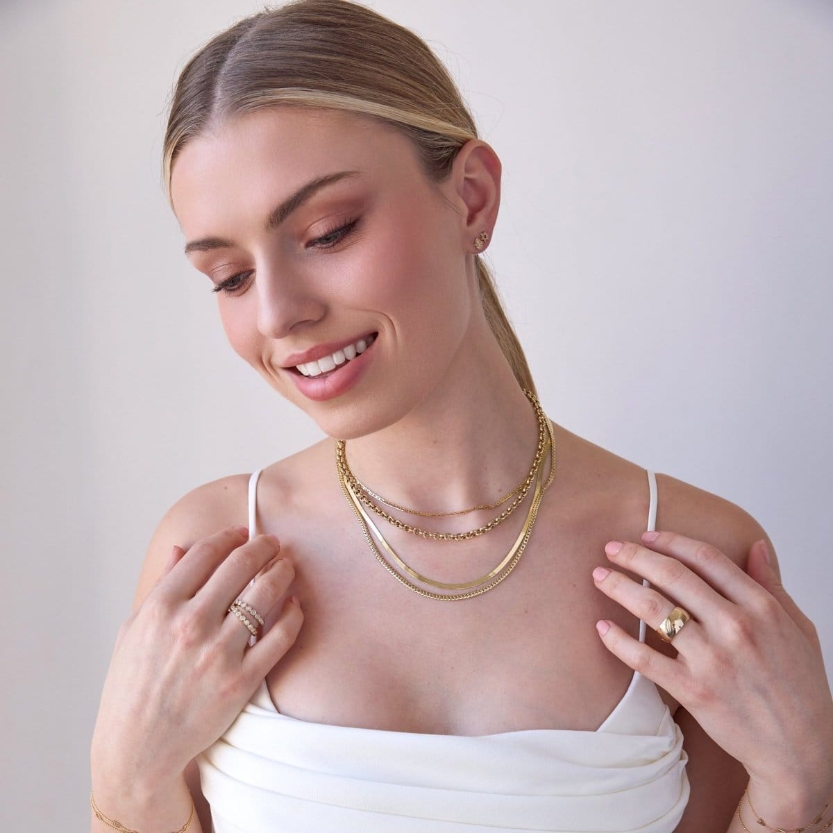 A blonde woman wearing layered yellow gold necklaces, including an Overboard Layering Chain, a Rolo Necklace, a Sequence Chain and a Chic Cuban Curb Chain, along with a Thick Band, two Evermore Rings, a Pavé Charm Stud - Hamsa and a Pavé Charm Stud - Star of David, with a white top against a plain background.