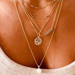 Close-up of a décolletage wearing several gold necklaces - an Element Short Link Chain, a Custom Gold Necklace - 2 Letters, a MAMA Necklace, a Zodiac Necklace - Capricorn and a Perla Necklace. 
