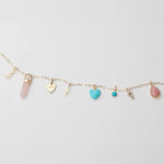 A yellow gold Element Short Link Chain with several pendants and charms, including Petite Nameplate Pendants in 'teddy', 'elise' and 'bijou', a rose quartz Balance Crystal Charm, an Engraved Heart Charm, an Etched Turquoise Heart Charm, a Turquoise Dangle Charm and a Pink Opal Tear Drop Charm, against a light grey background. 