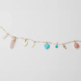 A yellow gold Element Short Link Chain with several pendants and charms, including Petite Nameplate Pendants in 'teddy', 'elise' and 'bijou', a rose quartz Balance Crystal Charm, an Engraved Heart Charm, an Etched Turquoise Heart Charm, a Turquoise Dangle Charm and a Pink Opal Tear Drop Charm, against a light grey background. 