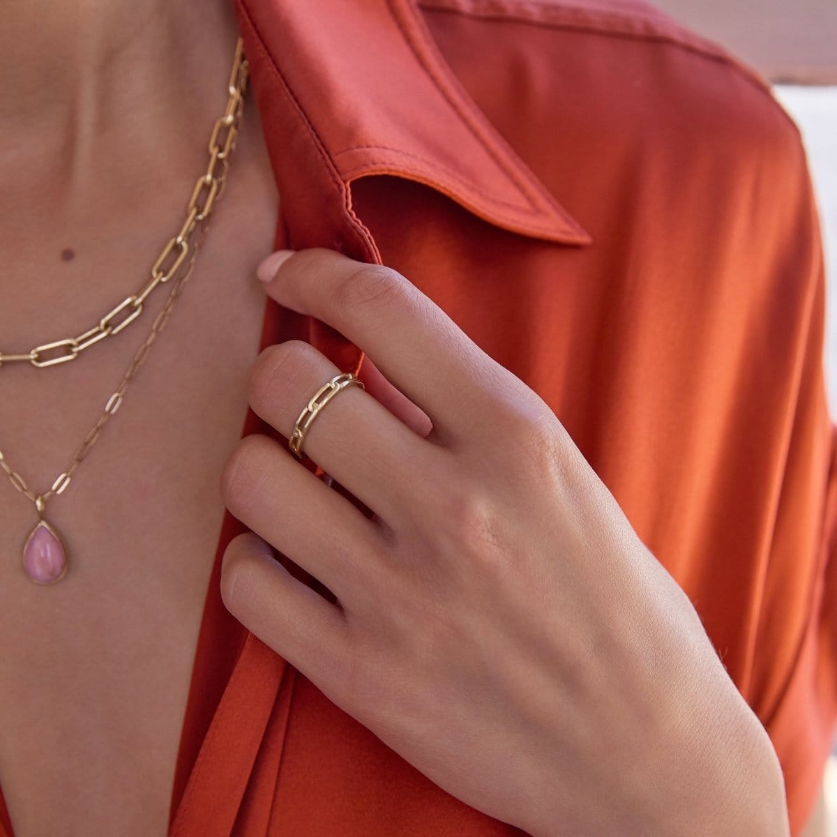 A close-up of a woman's décolletage and hand wearing a yellow gold Bold Link Chain, an Element Short Link Chain with a Pink Opal Tear Drop Charm and a Link Ring, with an orange shirt.