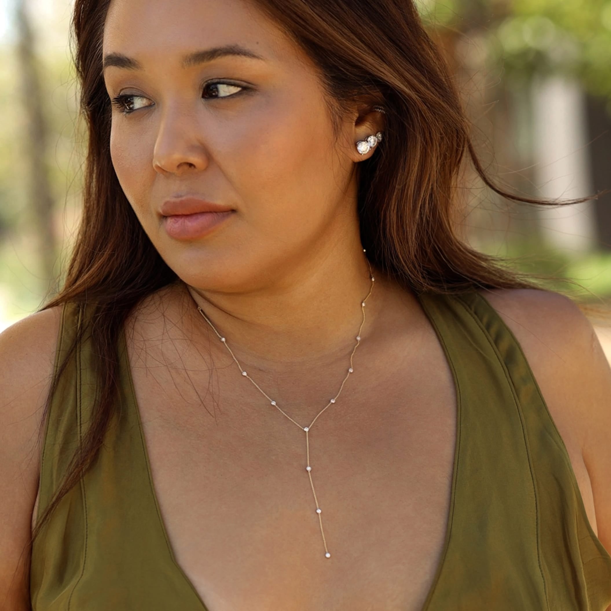 A woman wearing a delicate silver Y-necklace with small bead accents and a matching stud earring, photographed outdoors with a softly blurred natural background.