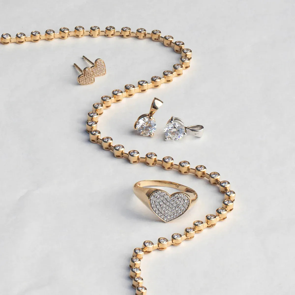 The Ultimate Diamond Necklace, a Love on the Rocks Ring, Pave Charm Studs -  Heart and two Diamond Pendants laid on top of a light grey surface. 