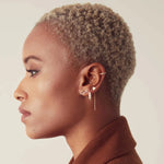 A More Love Stud, Tres Pearl Earring, 14K Gold Charm Threader Earring and Pave Double Ear Cuff worn by a woman with short blonde hair showing her side profile against a light grey background. 