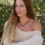 Woman with long blonde hair wearing a beige off-shoulder top and multiple necklaces against a natural background.