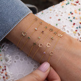 A close-up of a person's wrist wearing six delicate gold chain bracelets, each adorned with a different symbol or letter (such as a cross, crescent moon, star, and letters C and W) and small colorful gemstones. The person is wearing a denim sleeve and a white lace top.