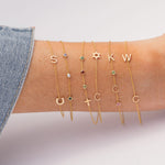 A close-up of a person's wrist wearing five delicate gold bracelets with letter charms and small gemstone accents, paired with a denim sleeve against a soft light gray background.