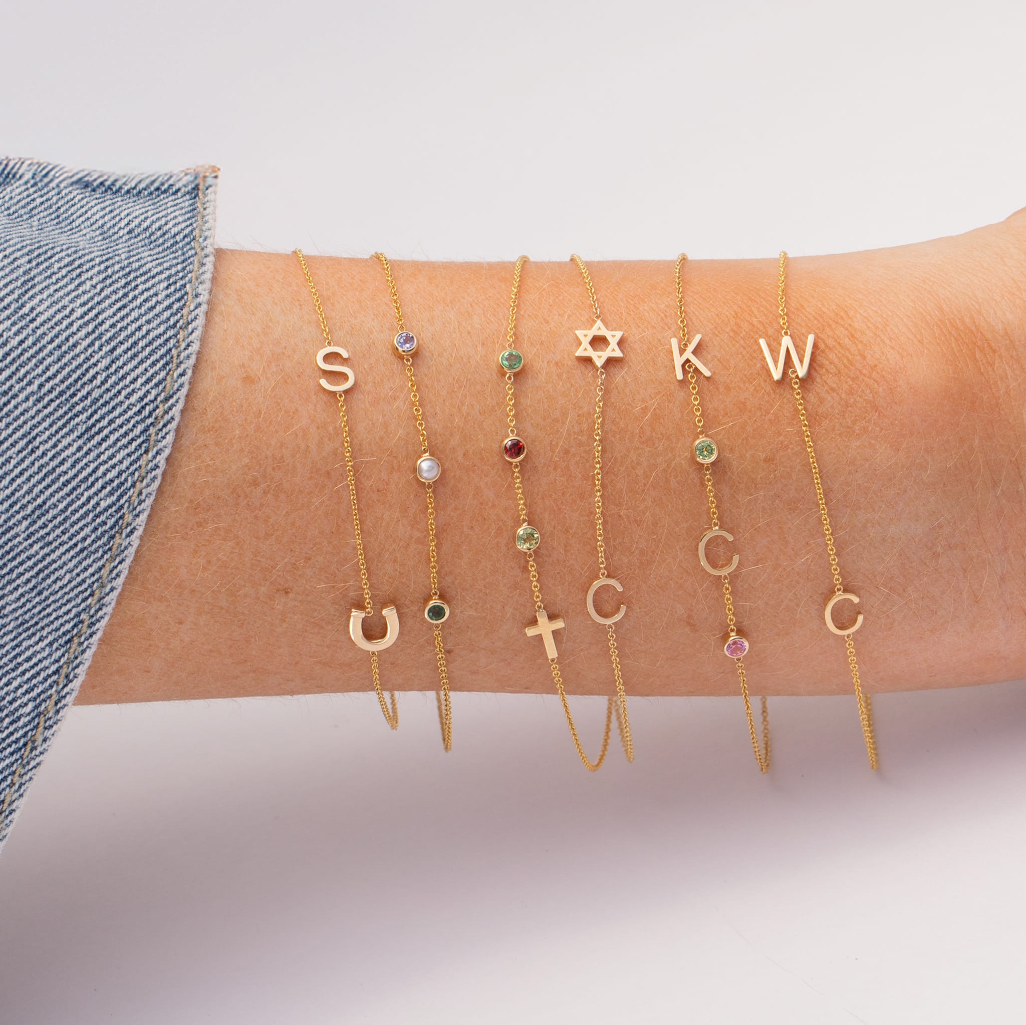 A close-up of a person's wrist wearing five delicate gold bracelets with letter charms and small gemstone accents, paired with a denim sleeve against a soft light gray background.