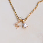 A gold chain necklace with a pendant featuring a clear round gemstone and a small gold letter 'M' embellished with tiny clear stones, shown on a light beige background.