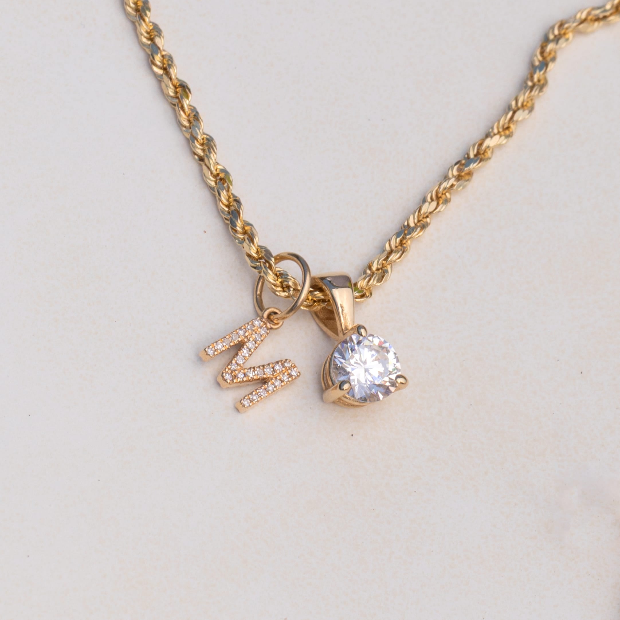 A gold chain necklace with a pendant featuring a clear round gemstone and a small gold letter 'M' embellished with tiny clear stones, shown on a light beige background.