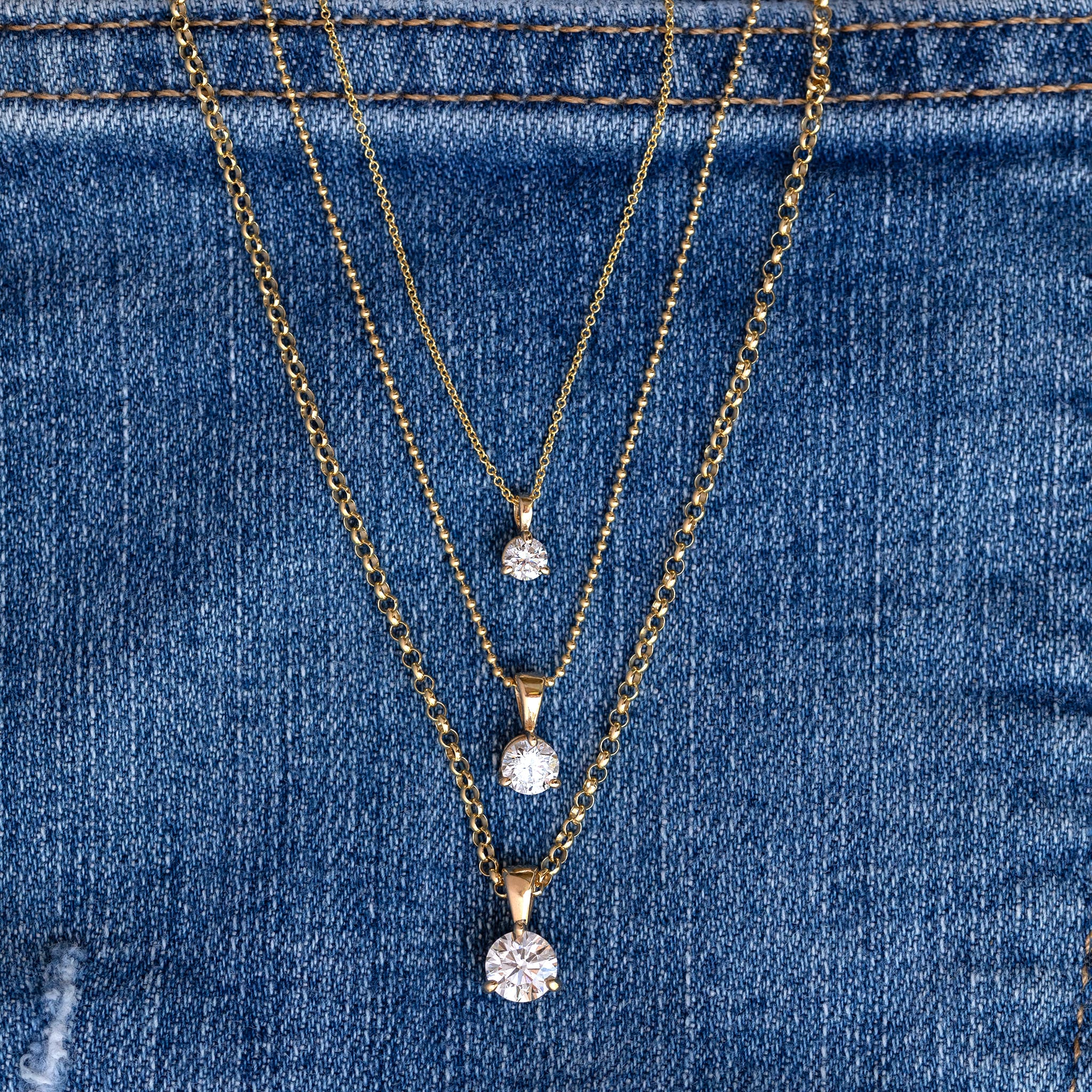 Three gold chain necklaces with round clear gemstone pendants in graduated sizes, displayed on a blue denim fabric background.
