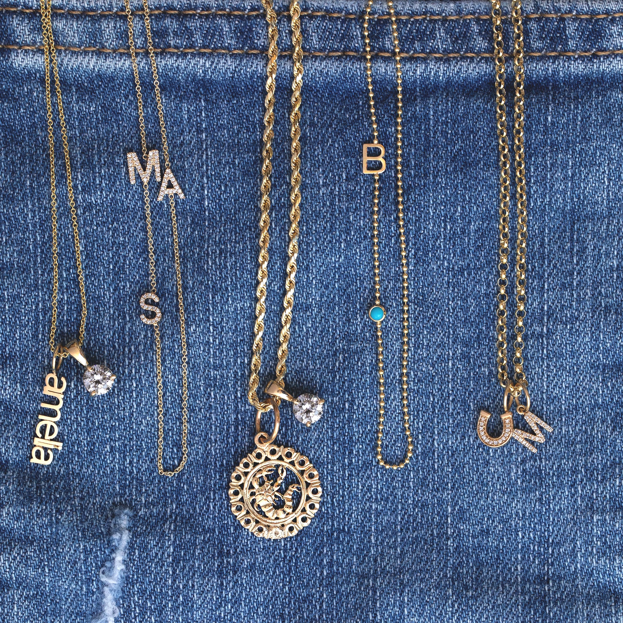 Five gold initial necklaces with letter charms are draped over a blue denim fabric background. The necklaces feature various letter designs, including 'MA', 'S', 'B', 'CM', and 'anlea', with some adorned with small clear rhinestones or a tiny turquoise stone.