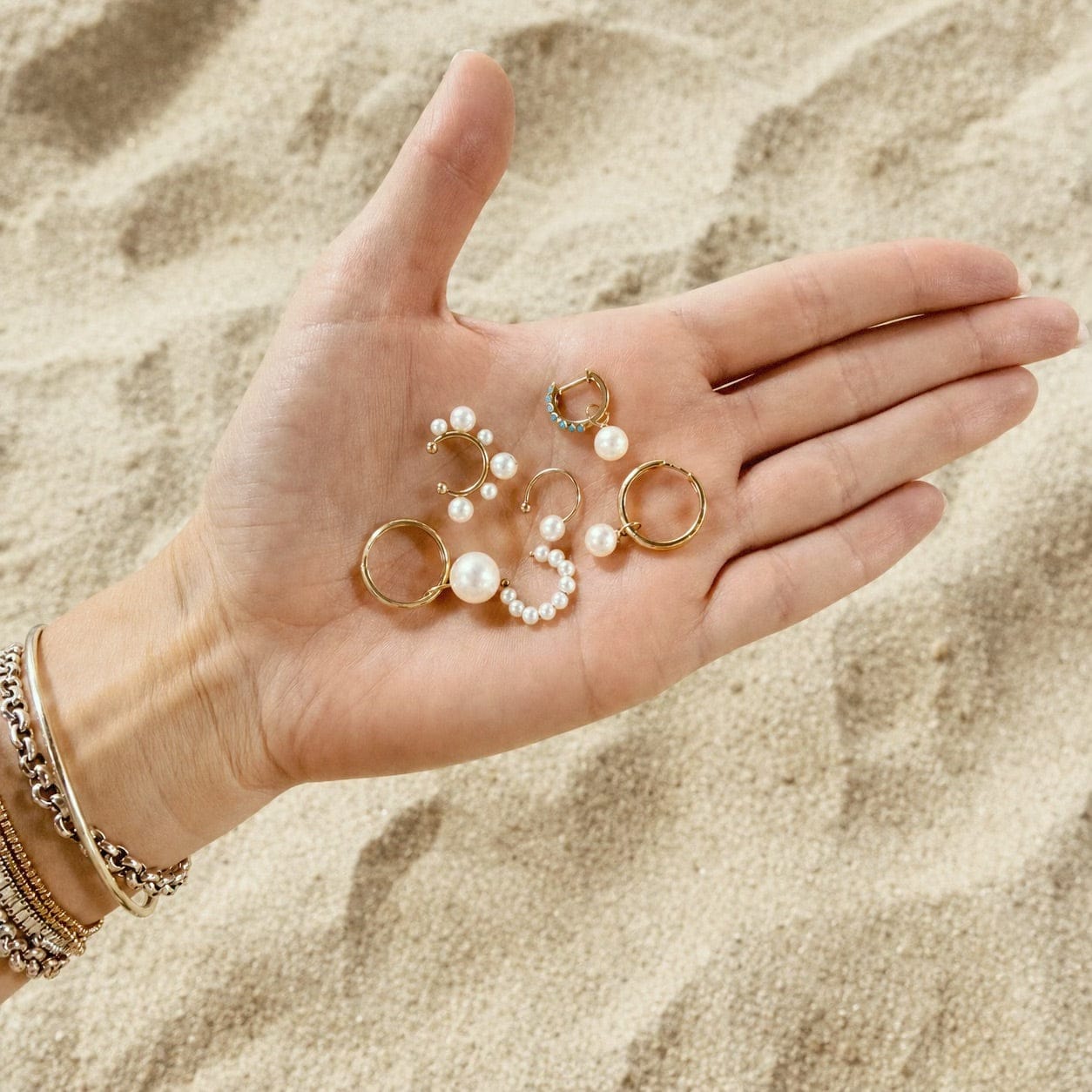 Maya Brenner pearl earring collection including Orbit Pearl Ear Cuff and pearl hoop earrings held in an open hand over sandy beach