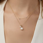 Gold necklace with a pearl pendant layred with a dainty initial necklace on a person wearing a white top.
