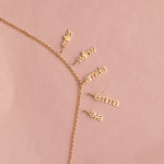 A delicate gold chain necklace laid flat on a dusty pink background, featuring five small dangling nameplates in cursive script reading 'miller', 'willow', 'amefa', 'emma', and 'ava'.