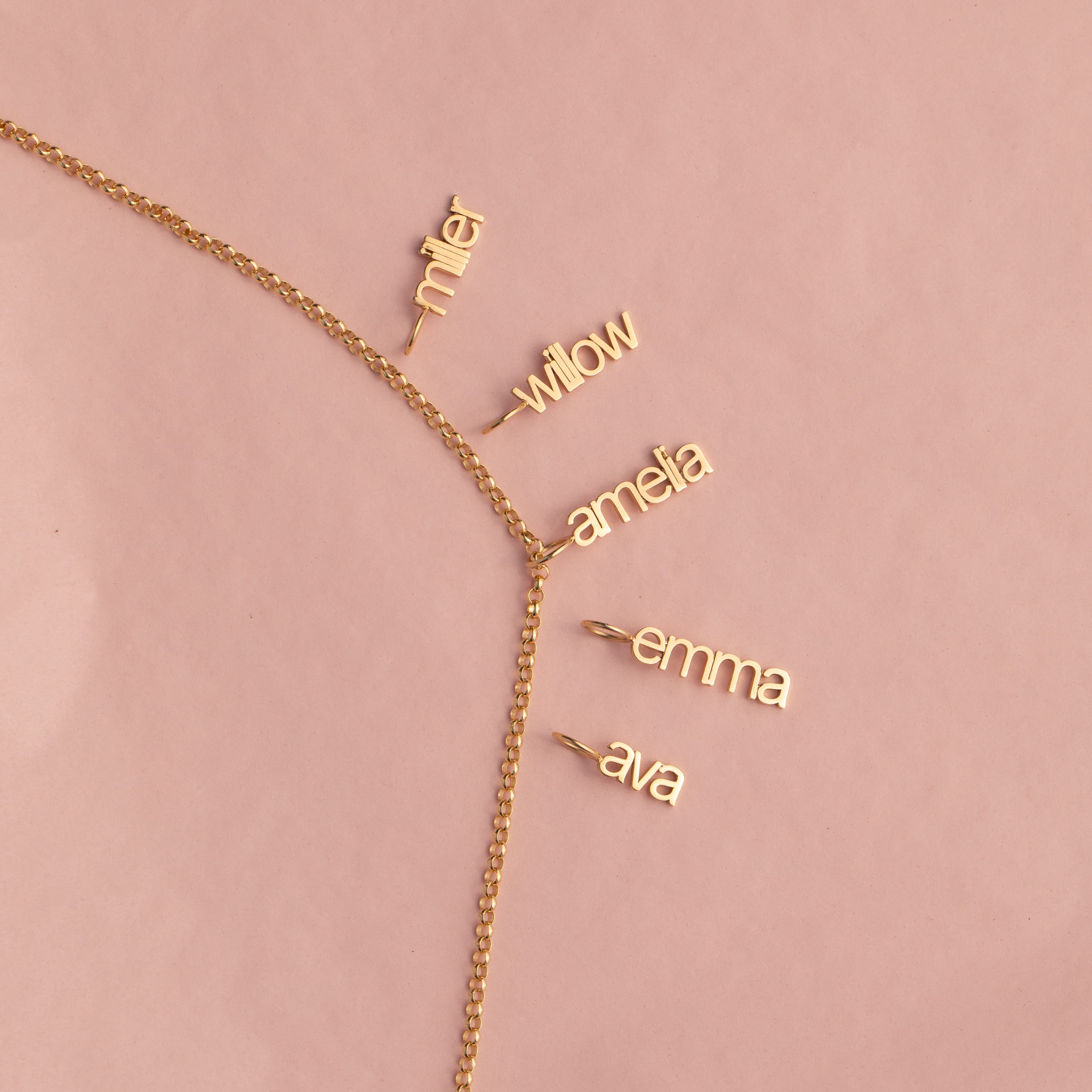 A delicate gold chain necklace laid flat on a dusty pink background, featuring five small dangling nameplates in cursive script reading 'miller', 'willow', 'amefa', 'emma', and 'ava'.
