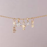 A gold chain necklace with three dangling nameplates spelling 'anabel', 'miller', and 'elva', alongside two clear round gemstones, shown against a solid light gray background.