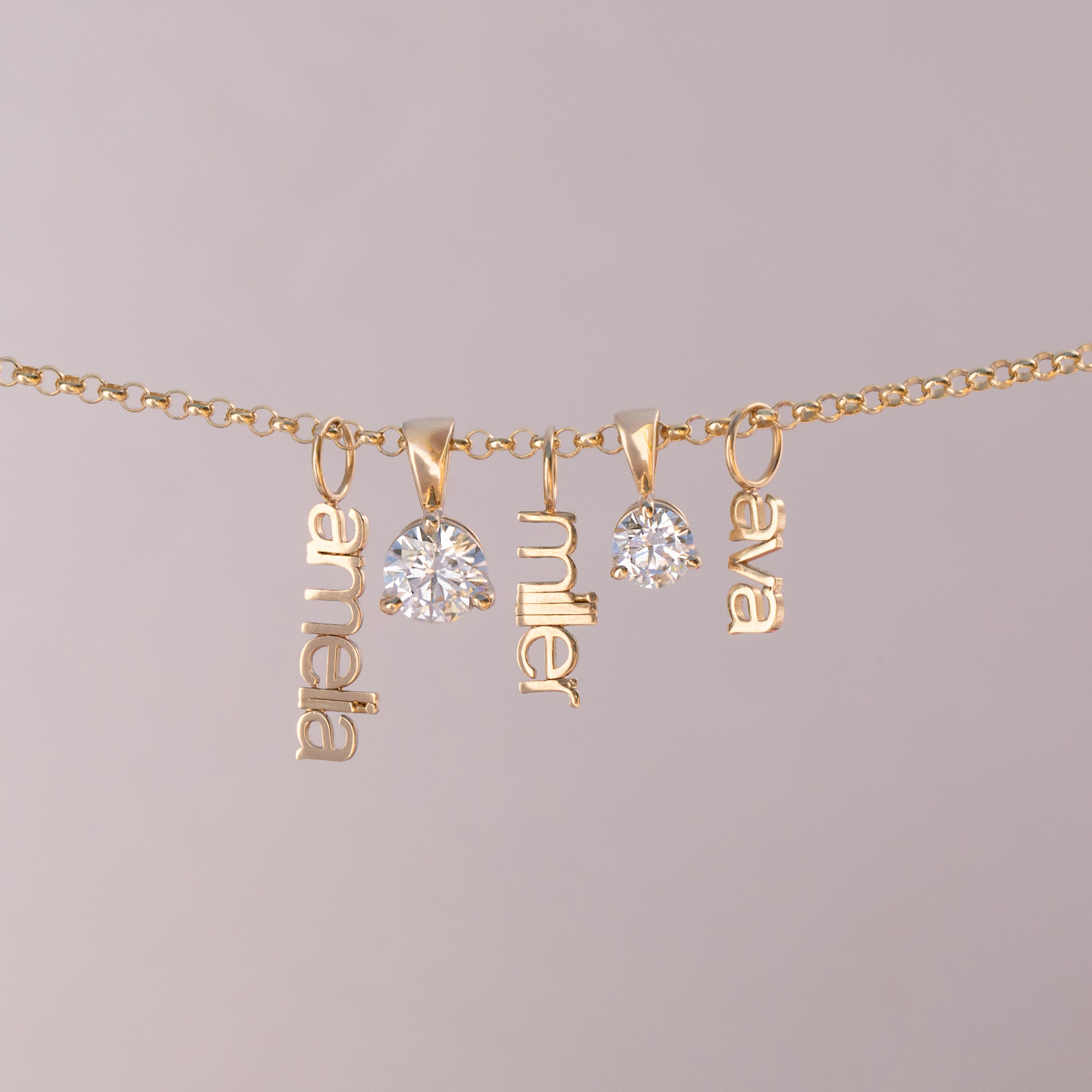 A gold chain necklace with three dangling nameplates spelling 'anabel', 'miller', and 'elva', alongside two clear round gemstones, shown against a solid light gray background.