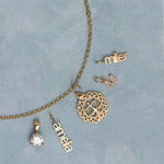 A gold jewelry set laid out on a textured light blue background, including a chain necklace with a circular scale of justice pendant, a solitaire diamond pendant, a nameplate reading 'anna', a nameplate reading 'miller', and a small nameplate reading 'M'.