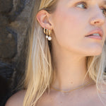 A close-up of a blonde woman wearing a pair of gold hoop earrings with small white pearl drops, along with a delicate gold chain necklace.