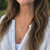 A close-up of a woman wearing layered gold necklaces, including one with a clear crystal point pendant, a small gray bead necklace, and a necklace with the word 'moai' in gold letters, styled with a white knit cardigan outdoors.