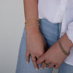 A close-up of a woman's hands wearing mixed metal jewellery, including a Classic Bubble Bracelet, Rolo Bracelets, a Diamond Cut Bracelet, Delicate Beaded Bands and a Thick Band, styled in a white shirt and light blue denim jeans. 