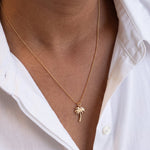 A close-up of a person wearing a delicate gold chain necklace with a small palm tree pendant, styled with an open white button-down shirt.