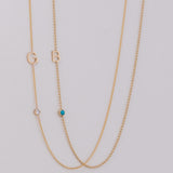 Two delicate gold chain necklaces laid flat on a light gray background. One necklace features a gold letter 'G' and a small clear stone, while the other features a gold letter 'B' and a small turquoise blue stone.