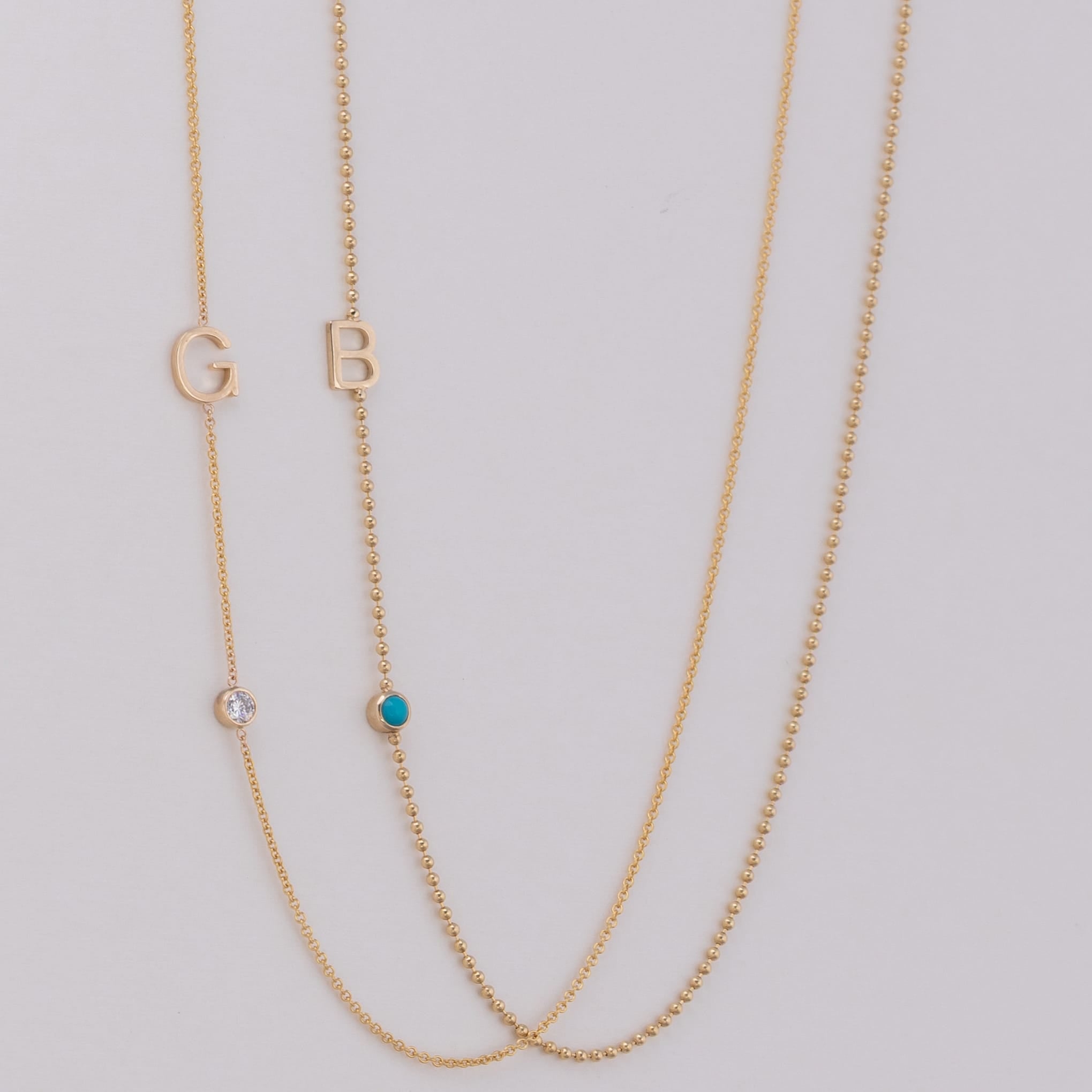 Two delicate gold chain necklaces laid flat on a light gray background. One necklace features a gold letter 'G' and a small clear stone, while the other features a gold letter 'B' and a small turquoise blue stone.