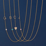 Five gold necklaces laid out on a dark blue background, each with a letter initial (G, B, C, J) and one necklace featuring a small heart pendant. The necklaces also have small round gemstone accents in clear, turquoise, and pink tones.