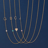Five gold necklaces laid out on a dark blue background, each with a letter initial (G, B, C, J) and one necklace featuring a small heart pendant. The necklaces also have small round gemstone accents in clear, turquoise, and pink tones.