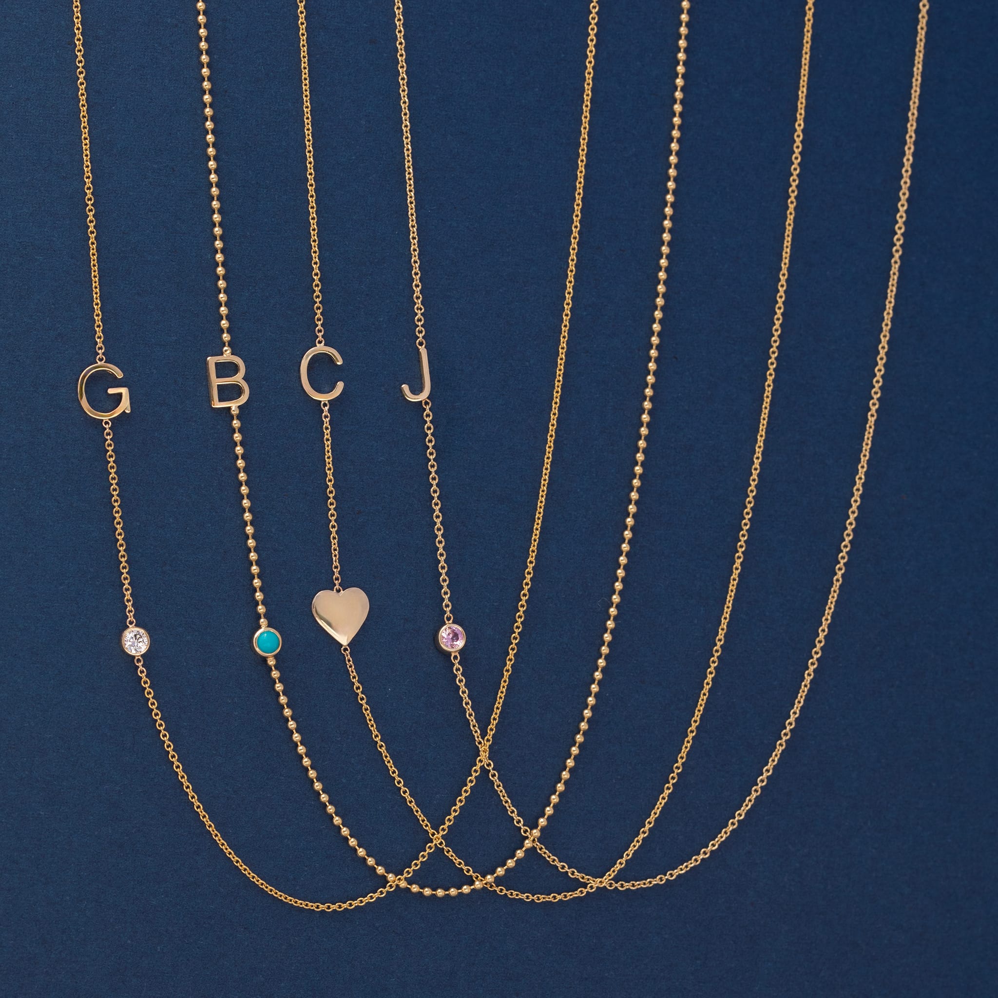 Five gold necklaces laid out on a dark blue background, each with a letter initial (G, B, C, J) and one necklace featuring a small heart pendant. The necklaces also have small round gemstone accents in clear, turquoise, and pink tones.