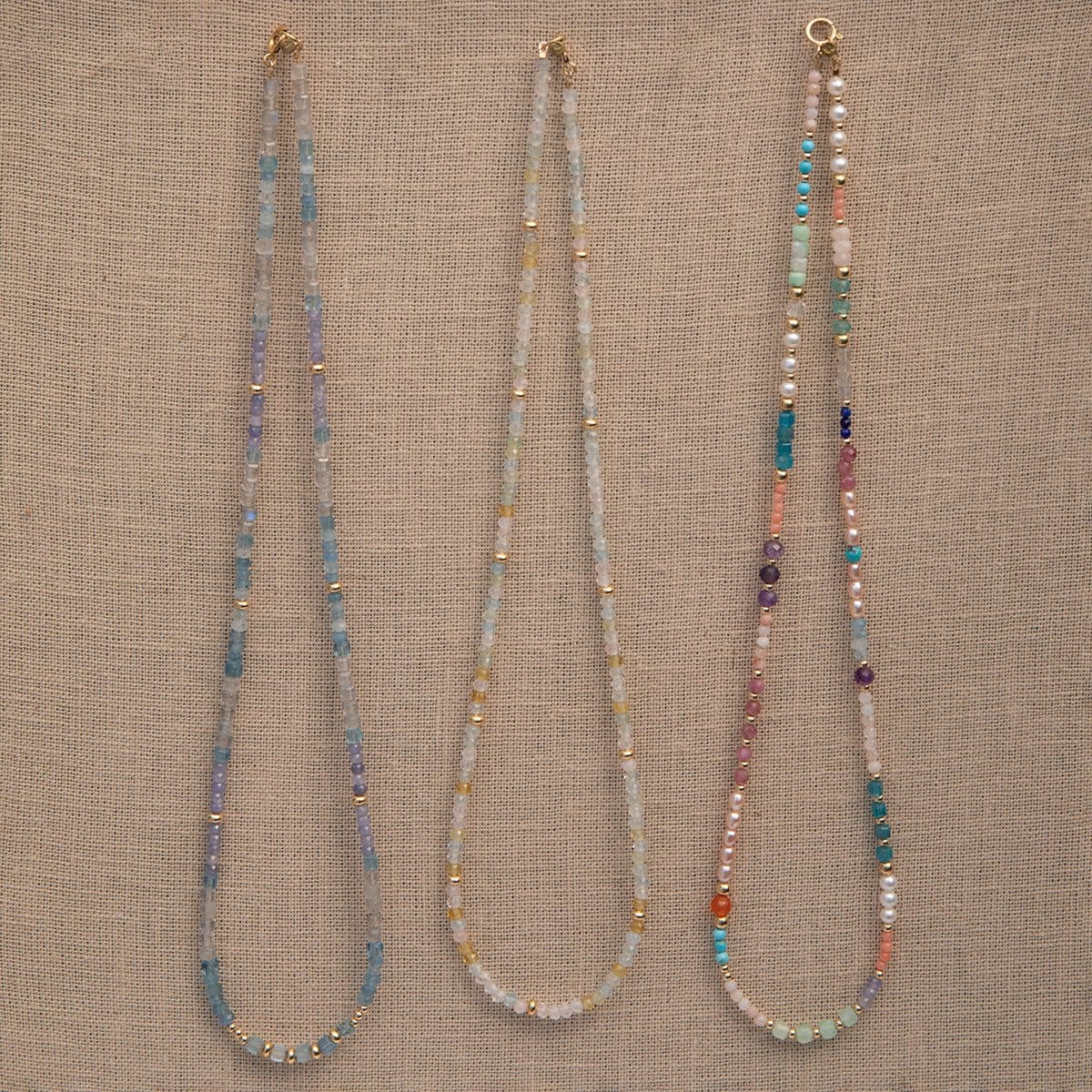 Three Endless Summer Necklaces (Blue Lagoon, Saint-Tropez and Capri) on a beige fabric background. 
